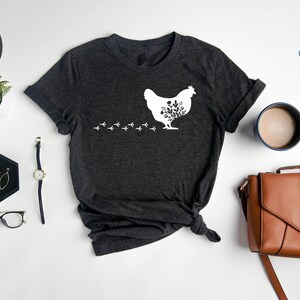 May include: Heather dark grey t-shirt with a white silhouette of a chicken with flowers inside. The chicken is walking to the left and leaving a trail of footprints behind it. The text "NFINITI Custom Shirts" is in the top right corner of the image.