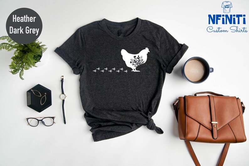 May include: Heather dark grey t-shirt with a white silhouette of a chicken with flowers inside. The chicken is walking to the left and leaving a trail of footprints behind it. The text "NFINITI Custom Shirts" is in the top right corner of the image.