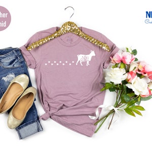 Pu&ograve; includere: Una t-shirt color orchidea heather con una silhouette bianca di una capra con dettagli floreali e una scia di impronte di zoccoli. La t-shirt &egrave; su un appendiabiti e sullo sfondo ci sono fiori e jeans.