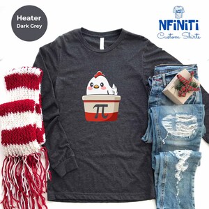 May include: Dark grey long-sleeve shirt with a cartoon chicken in a red and white container with the pi symbol. The shirt is next to a red and white scarf and blue jeans. The text "NFINiTi Custom Shirts" is in the upper right corner.