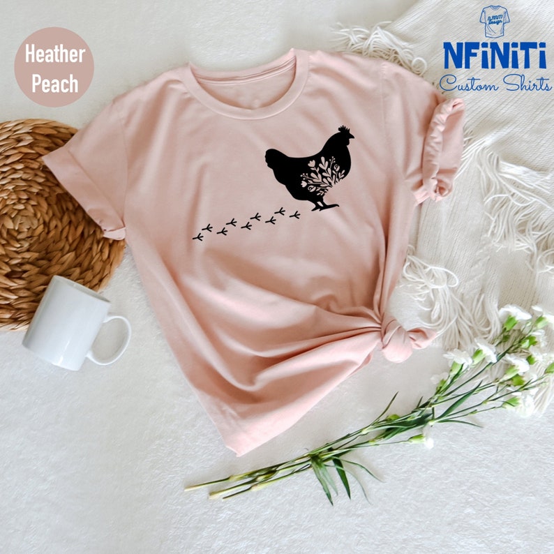 May include: A heather peach t-shirt with a black silhouette of a chicken with floral details and a trail of footprints. The text "NFINITI Custom Shirts" is in the top right corner of the image.