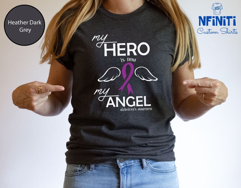 May include: Heather dark grey t-shirt with the text "my HERO is now my ANGEL" in white script. A purple ribbon and angel wings are featured in the design, with "alzheimer's awareness" below. The shirt is a custom design.