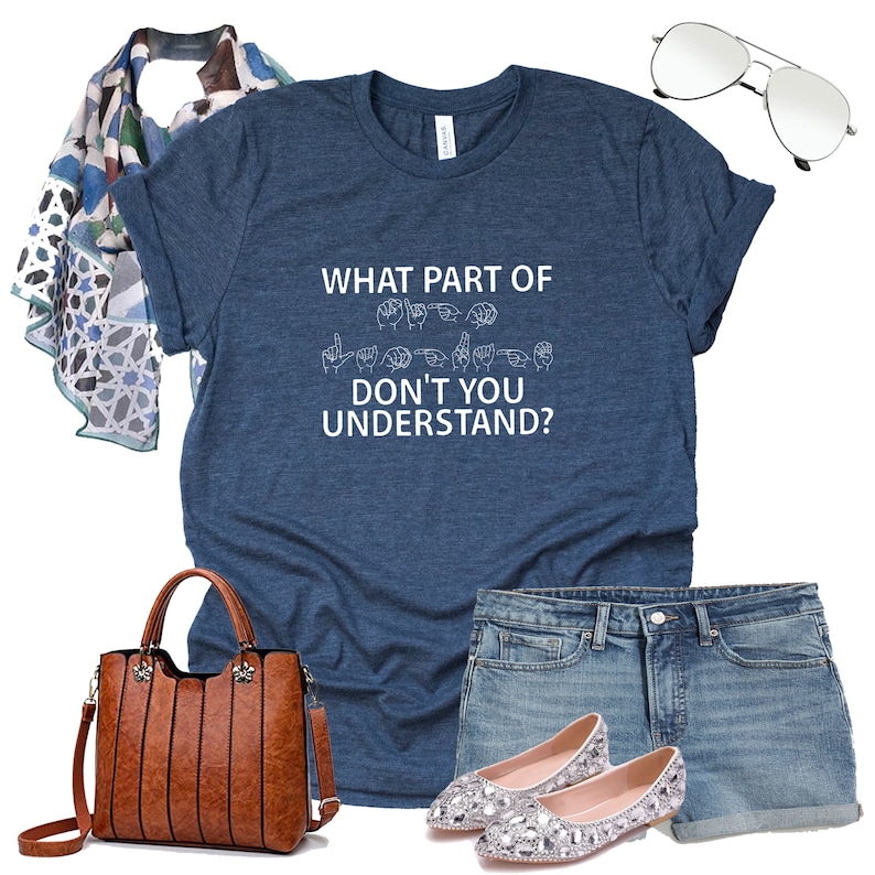 May include: A blue t-shirt with the text "WHAT PART OF DON'T YOU UNDERSTAND?" in white lettering. The text is accompanied by a row of hand signs in sign language. The shirt is paired with denim shorts, a brown leather handbag, and sparkly silver flats.