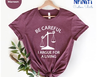 May include: Heather maroon t-shirt with white text that reads "Be Careful I Argue For A Living" and a white scale graphic.