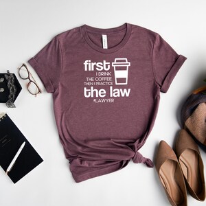 以下が含まれることがあります： ヘザーマロンのTシャツに、白い文字で「first I drink the coffee, then I practice the law #lawyer」と書いてあります。Tシャツは裾が結ばれています。Tシャツは白い背景に他のアクセサリーと一緒に撮影されています。