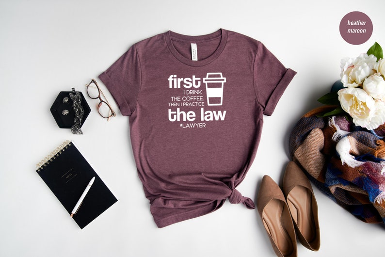 以下が含まれることがあります： ヘザーマロンのTシャツに、白い文字で「first I drink the coffee, then I practice the law #lawyer」と書いてあります。Tシャツは裾が結ばれています。Tシャツは白い背景に他のアクセサリーと一緒に撮影されています。