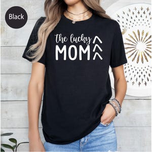 May include: Black t-shirt with the text "the lucky MOM" in white, with two upward-pointing arrows. The shirt is short-sleeved and appears to be made of a soft material. The word "Black" is in a black circle in the upper left corner.