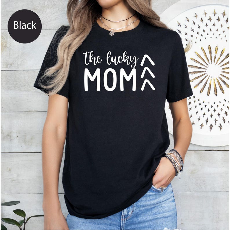 May include: Black t-shirt with the text "the lucky MOM" in white, with two upward-pointing arrows. The shirt is short-sleeved and appears to be made of a soft material. The word "Black" is in a black circle in the upper left corner.
