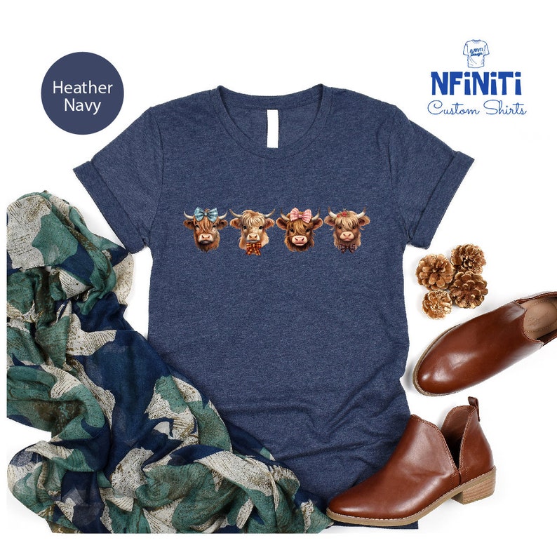 Puede incluir: Camiseta azul marino jaspeada con un gr&aacute;fico de cuatro vacas de las tierras altas con lazos. Las vacas son marrones con marcas blancas y tienen lazos de diferentes colores. El texto "NFINITI Custom Shirts" est&aacute; en la esquina superior derecha de la imagen.