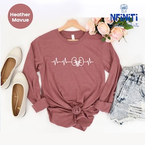 May include: A heather mauve long-sleeved t-shirt with a white graphic of a heartbeat with kidneys in the center. The shirt is on a white background with a pair of white flats and a pair of denim shorts.