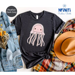 May include: A heather dark grey t-shirt with a cute cartoon pink jellyfish with a smiling face. The shirt is folded and displayed on a plaid fabric with a brown hat and a denim jacket.