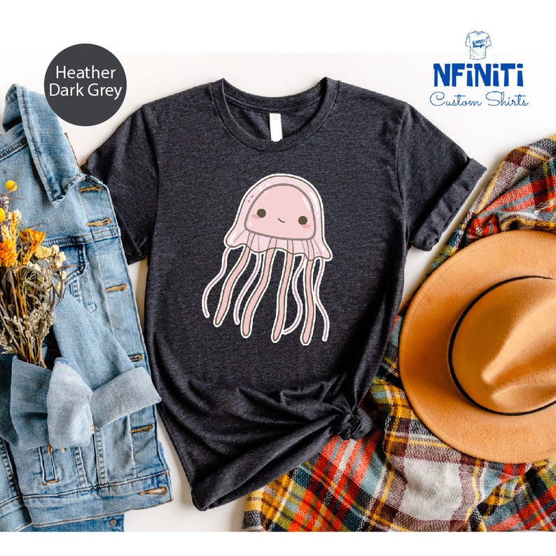 May include: A heather dark grey t-shirt with a cute cartoon pink jellyfish with a smiling face. The shirt is folded and displayed on a plaid fabric with a brown hat and a denim jacket.