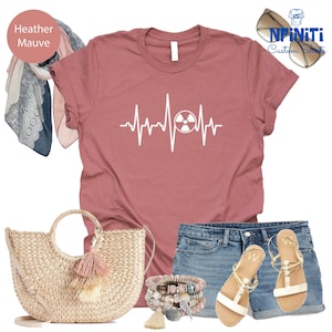 May include: Heather mauve t-shirt with a white graphic of a heartbeat with a radiation symbol inside. The shirt is paired with denim shorts, a straw bag with a tassel, and white sandals.