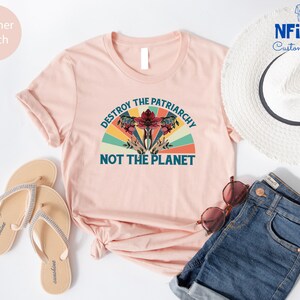 May include: A pink t-shirt with a vintage-style graphic that says "Destroy the Patriarchy Not the Planet". The graphic features a stylized flower with a red center and green leaves, surrounded by a rainbow-colored arc.