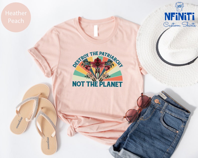 May include: A pink t-shirt with a vintage-style graphic that says "Destroy the Patriarchy Not the Planet". The graphic features a stylized flower with a red center and green leaves, surrounded by a rainbow-colored arc.