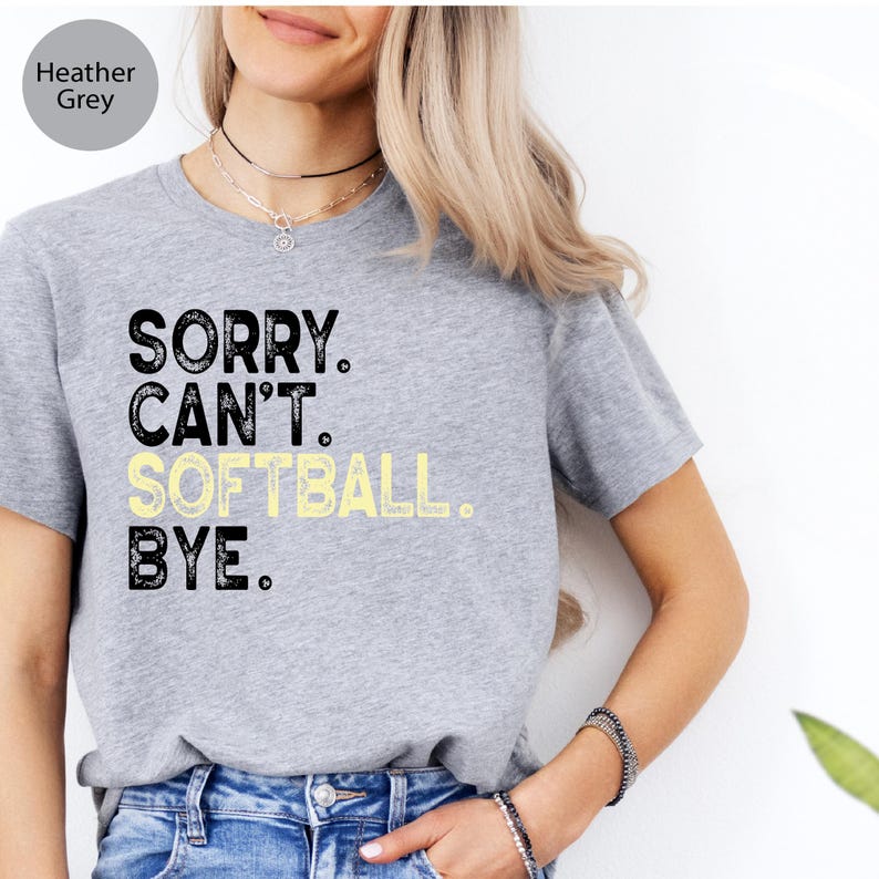 May include: Heather grey t-shirt featuring the text "SORRY. CAN'T. SOFTBALL. BYE." in black and yellow. The shirt has a crew neck and is made of a soft fabric.