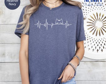 Può includere: T-shirt blu navy melange con grafica bianca di un volto di gatto integrato in una linea di battito cardiaco. La maglietta ha maniche corte e girocollo. Il testo "Heather Navy" è in un cerchio in alto a sinistra.