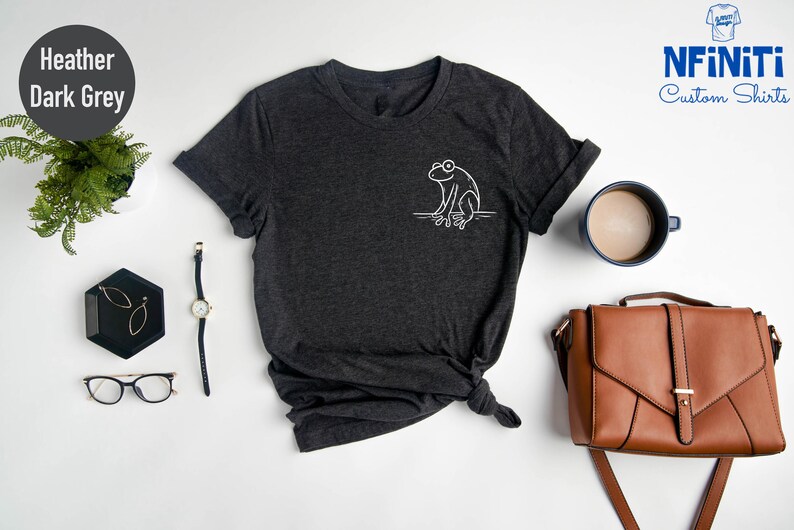 May include: Heather dark grey V-neck t-shirt with a white outline of a frog sitting on a branch. The shirt is laid flat on a white surface with a brown leather purse, a cup of coffee, a watch, a pair of glasses, and a small black tray with gold earrings.