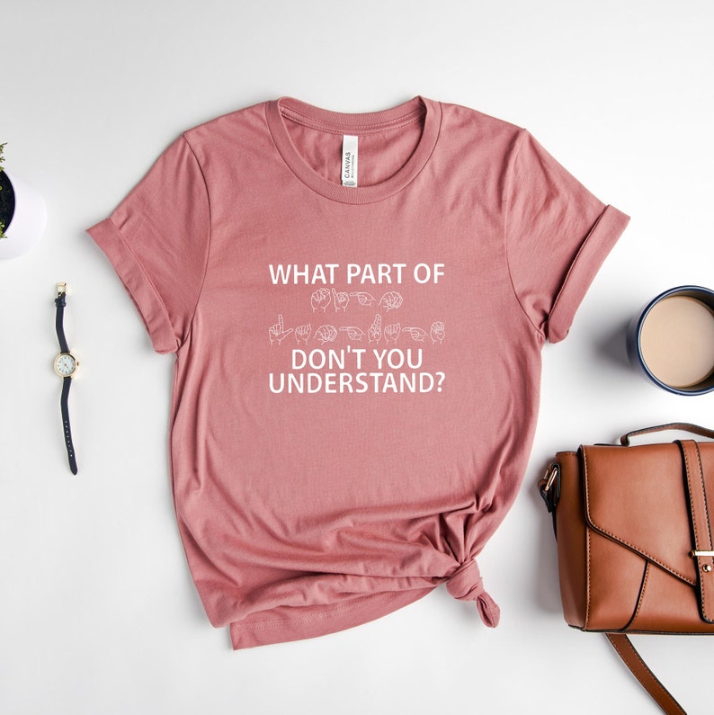 May include: A dusty rose t-shirt with the text "WHAT PART OF DON'T YOU UNDERSTAND?" in white. The text is accompanied by a row of white hand signs representing the phrase in American Sign Language.