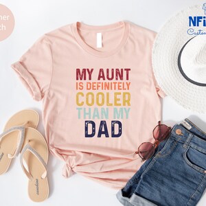 May include: A pink t-shirt with the text "My Aunt is Definitely Cooler Than My Dad" in a retro rainbow font.