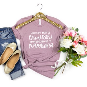 May include: A mauve t-shirt with the text "You people must be exhausted from watching me do everything" in white lettering. The t-shirt is on a gold hanger and is displayed with a pair of gold flats, blue jeans, and a bouquet of pink and white flowers.