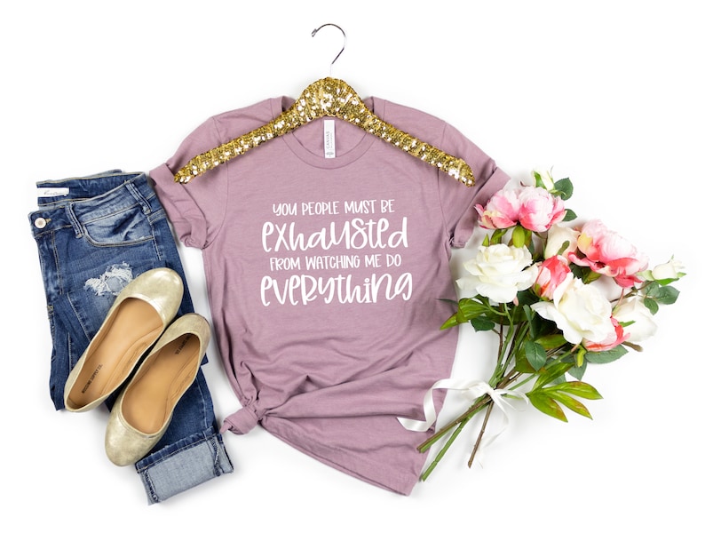 May include: A mauve t-shirt with the text "You people must be exhausted from watching me do everything" in white lettering. The t-shirt is on a gold hanger and is displayed with a pair of gold flats, blue jeans, and a bouquet of pink and white flowers.