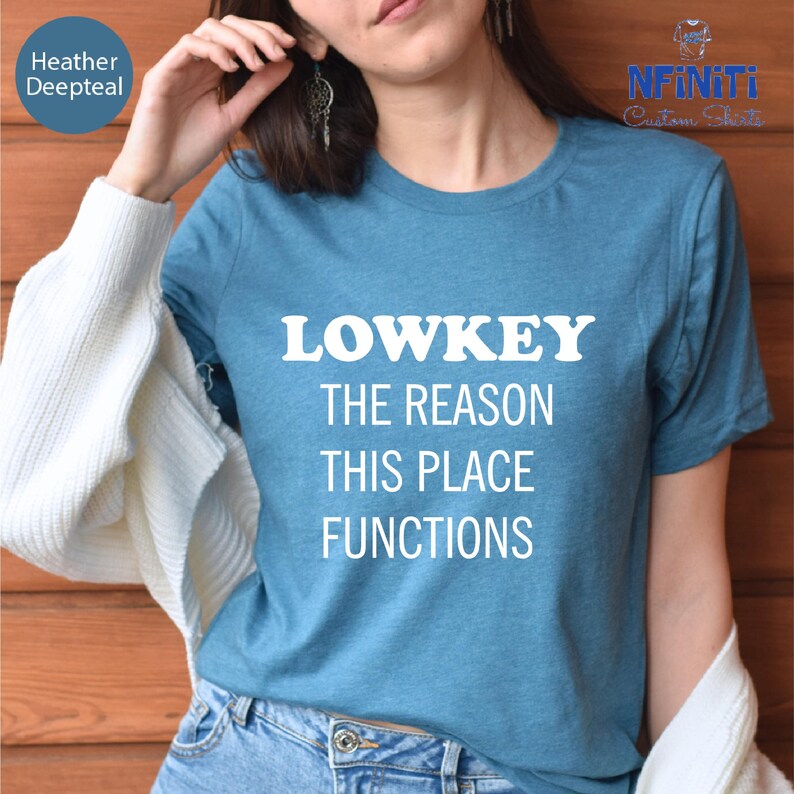 A woman wearing a blue t-shirt with the text "LOWKEY THE REASON THIS PLACE FUNCTIONS" printed on it.