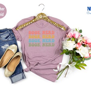 May include: A heather orchid t-shirt with the text "BOOK NERD" repeated four times in rainbow colors. The shirt is on a hanger and there are flowers in the background.