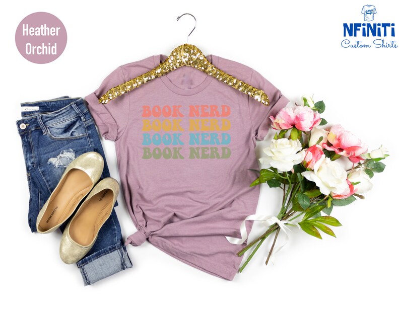 May include: A heather orchid t-shirt with the text "BOOK NERD" repeated four times in rainbow colors. The shirt is on a hanger and there are flowers in the background.
