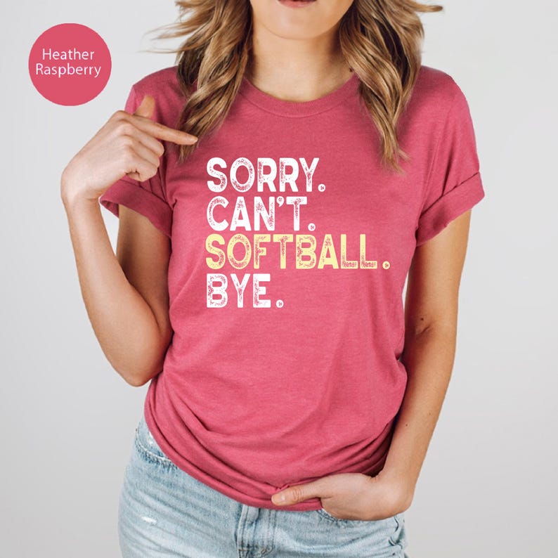 May include: A heather raspberry-coloured t-shirt featuring the text "SORRY. CAN'T. SOFTBALL. BYE." in white and yellow. The top has short sleeves and a crew neck.