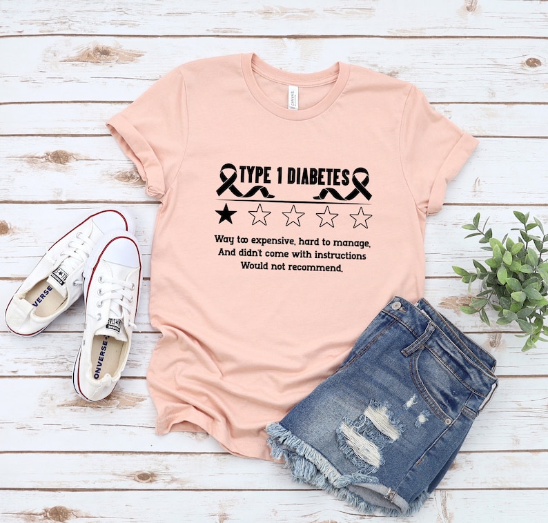 K&ouml;nnte beinhalten: Ein pinkfarbenes T-Shirt mit einer schwarzen Grafik, auf der "TYPE 1 DIABETES" mit einem schwarzen Band auf jeder Seite und einer 1-Sterne-Bewertung steht. Der Text unter der Grafik lautet "Way to expensive, hard to manage, And didn't come with instructions Would not recommend."