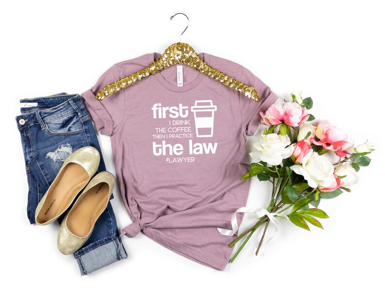 以下が含まれることがあります： コーヒーカップのグラフィックと「first I drink the coffee then I practice the law #lawyer」というテキストが書かれた、薄紫色のTシャツです。