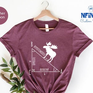 Può includere: Una t-shirt color marrone rossiccio con una grafica bianca di un alce in piedi su un triangolo rettangolo. Il triangolo è etichettato con le parole "ipotenusa", "opposto" e "adiacente".