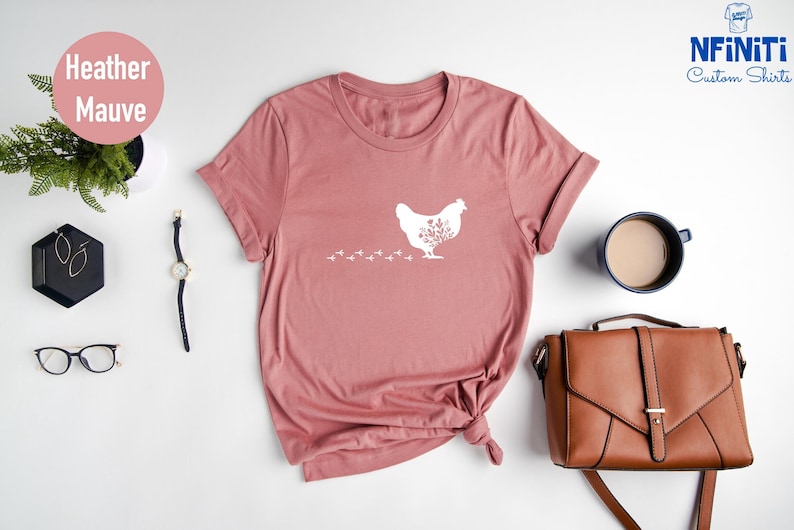 May include: Heather mauve t-shirt with a white graphic of a chicken with floral details. The chicken is walking with a trail of footprints behind it. The shirt is laid flat on a white surface.