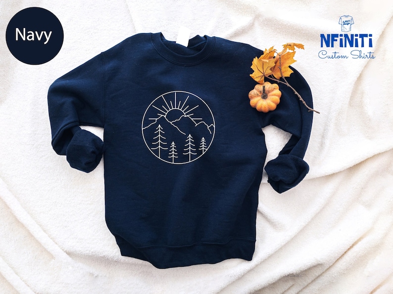 K&ouml;nnte beinhalten: Ein marineblauer Pullover mit einem wei&szlig;en Umriss einer Bergkette, Sonne und B&auml;umen in einem Kreis auf der Vorderseite. Der Pullover liegt auf einer wei&szlig;en, flauschigen Oberfl&auml;che.