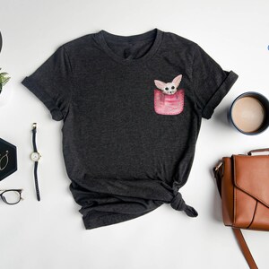 May include: Dark grey heather t-shirt with a pocket design featuring a cartoon fox. The shirt is styled with a watch, glasses, and a brown handbag. The brand name "NFINITI Custom Shirts" is visible.