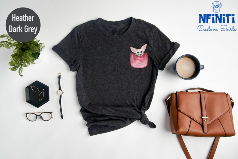 May include: Dark grey heather t-shirt with a pocket design featuring a cartoon fox. The shirt is styled with a watch, glasses, and a brown handbag. The brand name "NFINITI Custom Shirts" is visible.