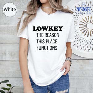 A woman wearing a white t-shirt with the text "LOWKEY THE REASON THIS PLACE FUNCTIONS" printed on it.