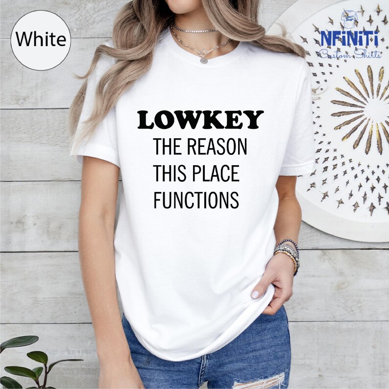 A woman wearing a white t-shirt with the text "LOWKEY THE REASON THIS PLACE FUNCTIONS" printed on it.