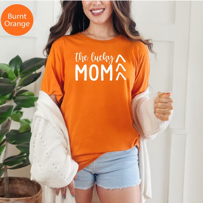 May include: Burnt orange t-shirt with the text "the lucky MOM" in white. The shirt is a crew neck style and is being worn with light blue shorts and a white cardigan. The shirt is a casual style.