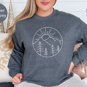 K&ouml;nnte beinhalten: Heather dunkelgrauer Pullover mit einem wei&szlig;en Umriss einer Sonne, Bergen und B&auml;umen in einem Kreis.