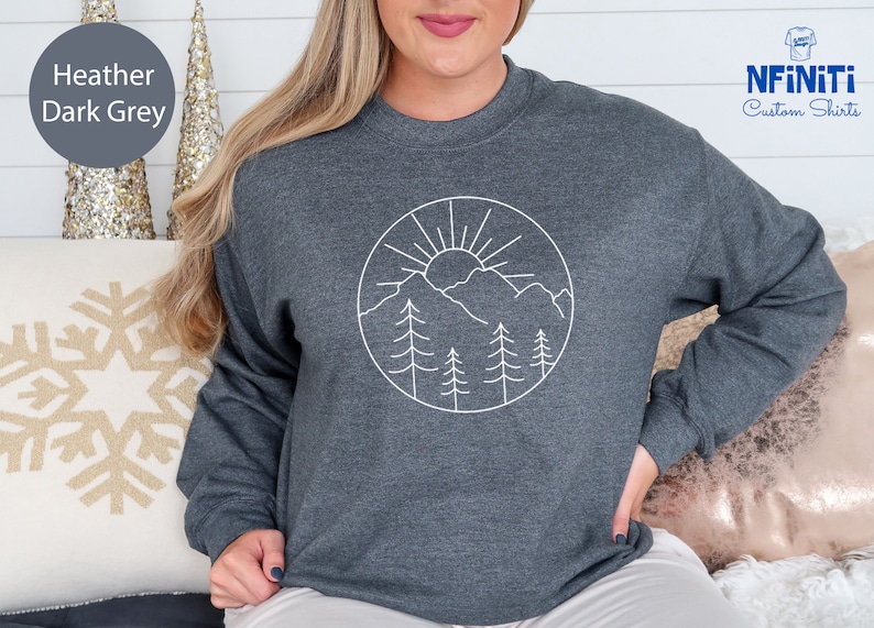 K&ouml;nnte beinhalten: Heather dunkelgrauer Pullover mit einem wei&szlig;en Umriss einer Sonne, Bergen und B&auml;umen in einem Kreis.