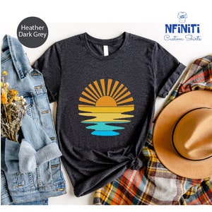 May include: A dark heather grey t-shirt with a graphic of a sunset over water. The graphic is in shades of orange, yellow, blue, and green. The text "NFINITI Custom Shirts" is in the top right corner of the image.