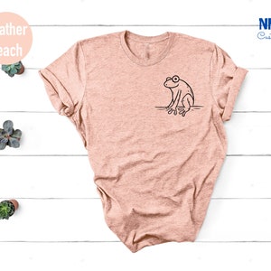 May include: A heather peach t-shirt with a black outline of a frog sitting on a branch. The shirt is on a white wooden background.