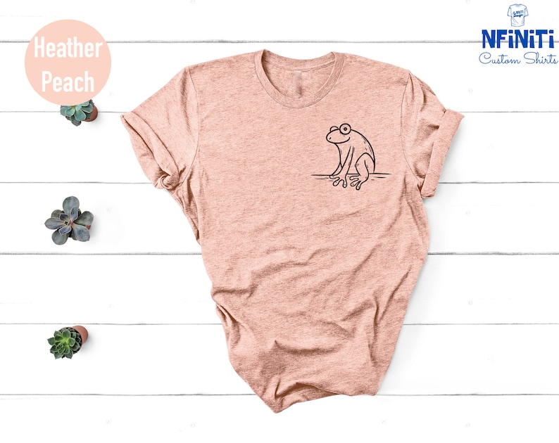 May include: A heather peach t-shirt with a black outline of a frog sitting on a branch. The shirt is on a white wooden background.