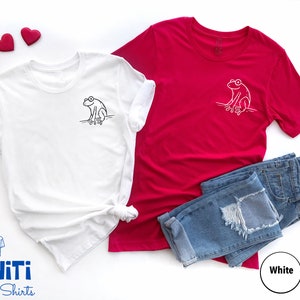 May include: Two t-shirts with a frog graphic design. One shirt is white with a black frog and the other is red with a white frog.