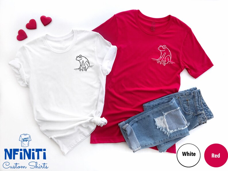May include: Two t-shirts with a frog graphic design. One shirt is white with a black frog and the other is red with a white frog.