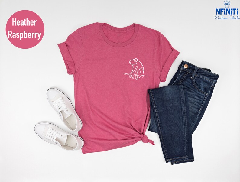 May include: A heather raspberry t-shirt with a white outline of a frog sitting on a branch. The shirt is shown on a white background with a pair of white sneakers and blue jeans.