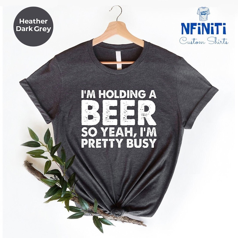 May include: A dark heather grey t-shirt with the text "I'm holding a beer so yeah, I'm pretty busy" printed in white.