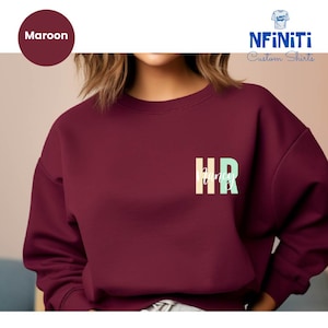 May include: A maroon sweatshirt with the text "HR" in a colorful block font and the word "Nancy" written in a white cursive font below it.